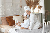 Loving mother take care to little daughter in white bathrobes and with towel on heads after a shower playing at morning on bed at cozy home