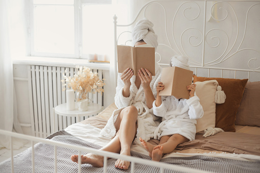 Mother and daughter in same bathrobes lying on the bed together and reading book at cozy home. Cover book mock up.