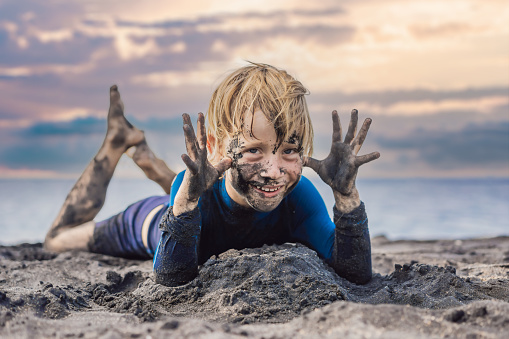 Black Friday concept. Smiling boy with dirty Black face sitting and playing on black sand beach. Summer vacation with kids. Black Friday, sales of tours and airline tickets or goods.
