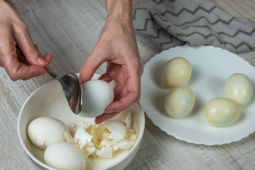 Removing the shell from boiled white eggs. A housewife takes an iron spoon and breaks the shell of a boiled chicken egg, peels the shell into a plate, puts the boiled egg into a bowl. Close-up.