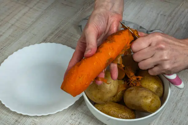 Removing the skin from boiled red carrots. Housewife with an iron kitchen knife peels a red-and-yellow carrot peel into a plate and puts the peeled carrots into a bowl. Concept of veganism. Close-up.