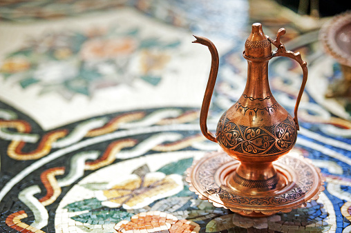 Jordan and the Arab world are known for their traditional crafts such as weaving, mosaics and brass and copper ware. Here a stylish traditional metal tea pot with curved spout and ornate design pattern sits on a mosaic surface in the souk.