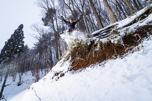 A Japanese snowboarder riding through backcountry forest drops off a small cliff section.