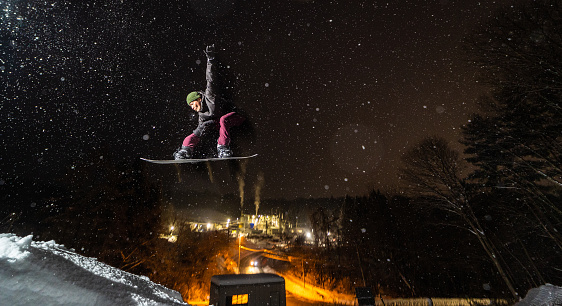 A Japanese man doing a snowboard jump at night with factory and street lights below while its snowing heavy.