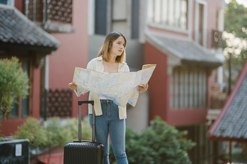 Young Asian woman solo traveler with suitcase using paper orientation map for walking while worried journey on the trip. Urban transportation and travel concept.