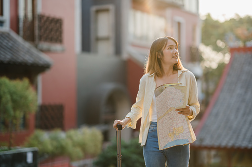 Young Asian woman solo traveler with suitcase using paper orientation map for walking around historic center. Urban transportation and travel concept.