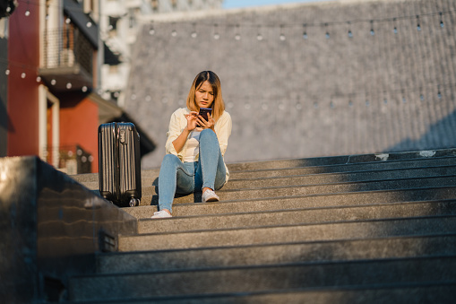 Young Asian woman solo traveler sitting on stairs with suitcase using phone gps while worried journey on the trip. Urban transportation and travel concept.