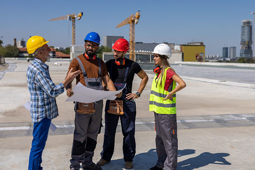 A Multicultural Group of Workers with Protective Helmets is Analyzing Blueprints at the Construction Site.