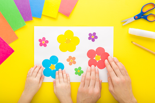 Young adult mother and child hands modeling colorful flower shapes from colorful paper on white sheet. Bright yellow table background. Playing and spending time together. Closeup. Top down view.