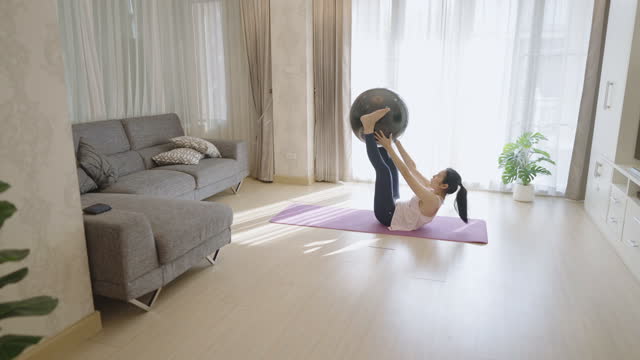 Asian woman exercising using fitness ball to do sit-ups by holding it with both hands over her head and sending it back and forth to her feet while lying on yoga mat at home. Home fitness, home workout.