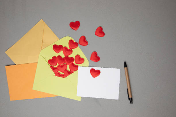 Light green, orange, yellow envelopes and a white card for text with red falling hearts on a gray concrete background. Happy Valentine's Day greeting, background. Flat lay