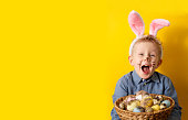 Cute boy with bunny ears holding basket with easter eggs on yellow background copy space