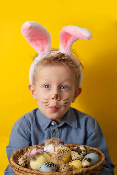 Cute boy with bunny ears holding basket with easter eggs on yellow background copy space