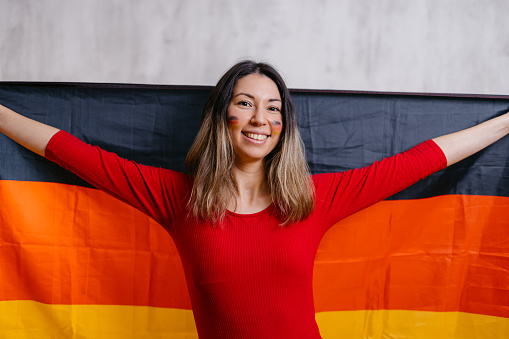 Portrait of a beautiful young woman with painted German flag colors on her cheeks indoors, holding up a German flag.