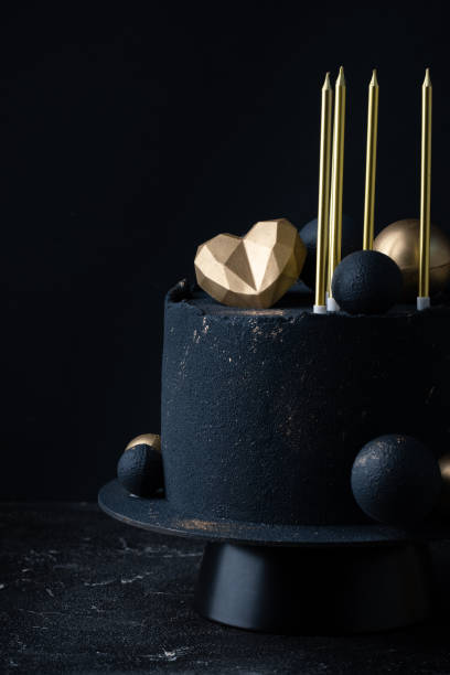 istock Luxury anniversary cake with black velvet sprayed coating decorated with golden chocolate spheres, heart and candles on top. Vintage cake for beloved one on the black background 1470938757