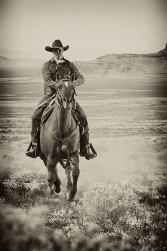 A man wearing old western attire rides a horse across a country plain. Vertical shot.