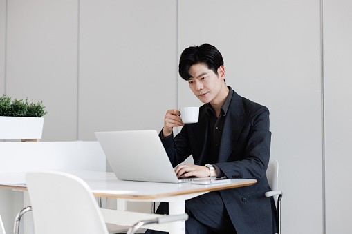 This is a young Asian male businessman, wearing a formal suit, sitting in a bright coffee shop, using a laptop to work remotely.
