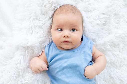 portrait of a cute baby boy three months old in a blue bodysuit on a white bed at home.