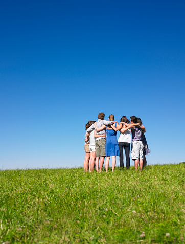 A group of people stands huddled together, with backs to the camera, on a grassy slope under blue sky. Vertical shot.