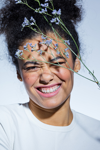Happy young woman wearing white blouse posing with delicate flowers next to her face, looking at camera and laughing. Close up of face.
