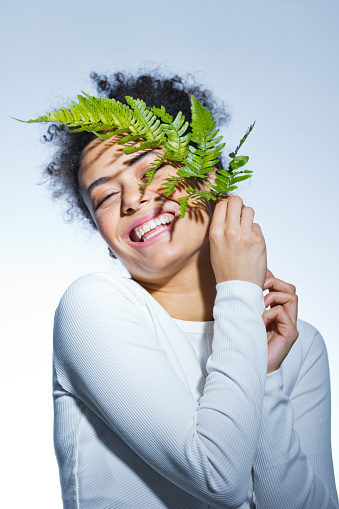 Beauty portrait of young woman wearing white blouse holding fern leaf next to her face, smiling with eyes closed.