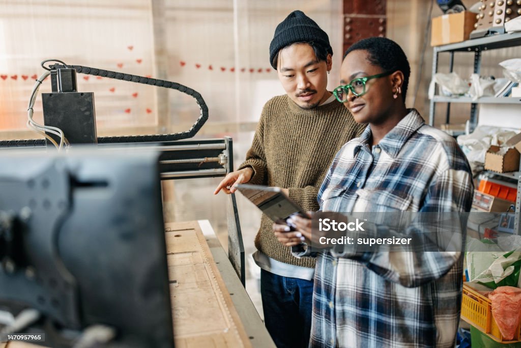 Innovating with Technology: Two Students Mastering the CNC Machine Two University Students with Different Ethnicities Operating CNC Machine with Digital Tablet 20-24 Years Stock Photo