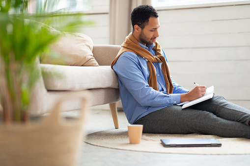 Business And Education Concept. Smiling young Arabic man sitting on floor leaning on couch writing letter in paper notebook, free copy space. Stylish millennial male studying, using pc, making plans