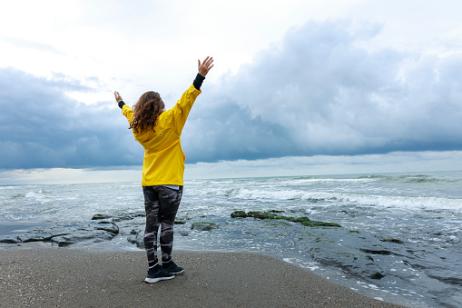 Young woman in yellow raincoat raising her hands against the sea in a stormy weather and feeling freedom