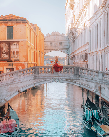 In this photo, a young woman wearing a stunning red dress is sitting on the Bridge of Sighs in St. Mark's Square in Venice, Italy. She is enjoying a delicious slice of pizza while taking in the beautiful views around her. The sun shines bright in the clear blue sky, casting a warm glow over the ancient bridge and the surrounding buildings.\nThe woman appears to be completely absorbed at the moment, savoring each bite of the mouth-watering pizza and soaking in the unique ambiance of Venezia Italia. Behind her, we can see the intricate architecture of the city's famous buildings and the shimmering waters of the canal below.\nOverall, this photo captures a beautiful moment of relaxation and indulgence in one of the most picturesque and iconic locations in Venice.