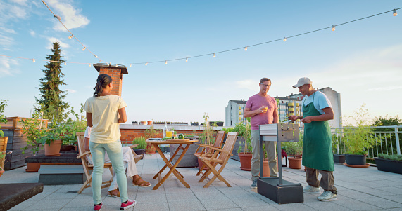 Group of friends having dinner and preparing food on barbecue grilled on rooftop garden.