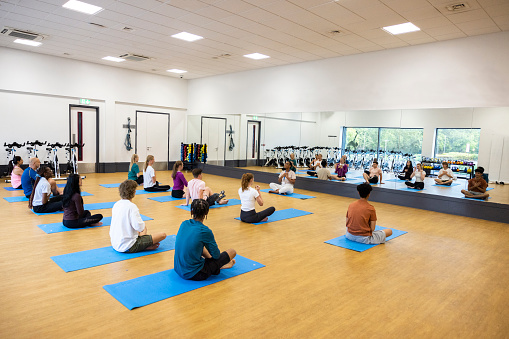 A wide-angle shot of a diverse group of teenage students participating in an indoor yoga class. They are sitting down on yoga mats and listening to the Yoga teacher. The yoga teacher is at the front of the class facing the students.