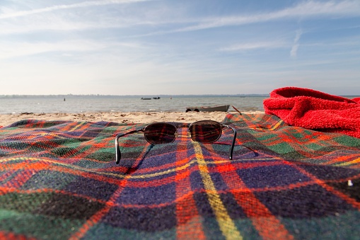 A pair of sunglasses lays on a beach blanket featuring a classic plaid pattern, with a red towel draped nearby