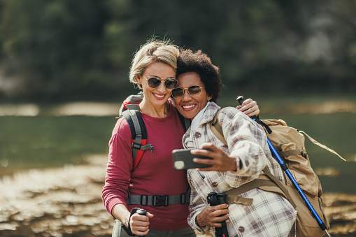 Two mixed-race female friends taking selfies by a river during a hike along the mountain.