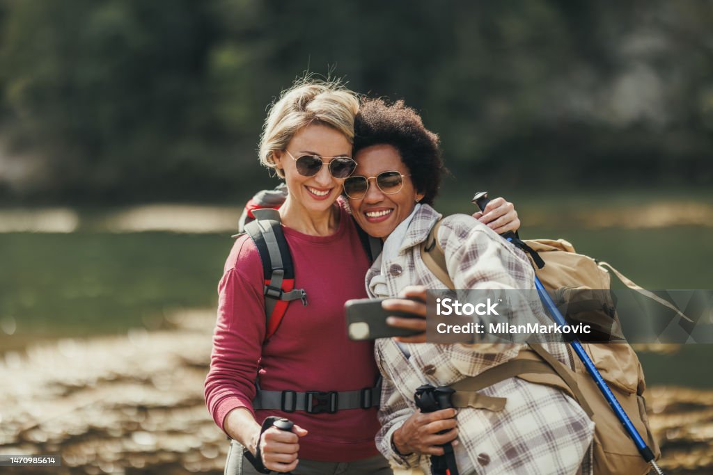 Two Women Enjoy Nature And Taking Video Call While Hike Two mixed-race female friends taking selfies by a river during a hike along the mountain. Hiking Stock Photo