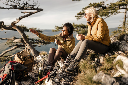 Two mixed-race female friends sitting on the top a hill and having tea break during her hike in the mountains in a beautiful sunny autumn day.
