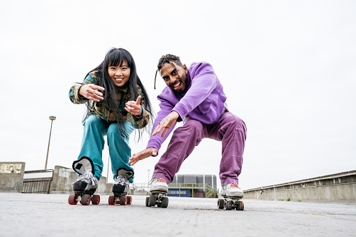 Low angle view of late 30s Asian woman and late 20s Black man, both in casual winter clothing, crouching, gesturing, and grinning as they approach camera.