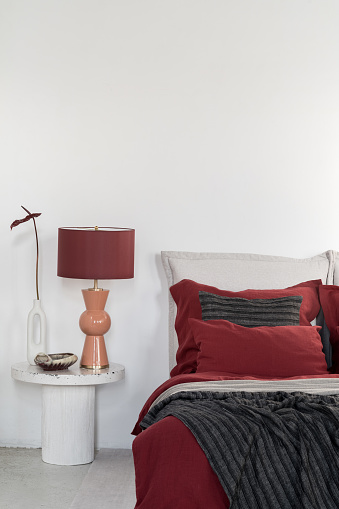 Elegant orange and red lamp and vase with flower on marble nightstand next to comfortable bed with red and black bedclothes and pillows in bright bedroom