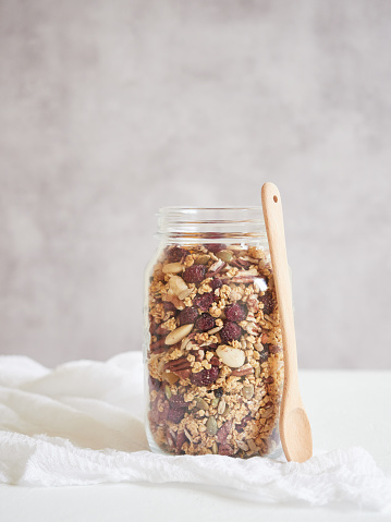Glass jar filled with delicious healthy nuts and dried fruits placed on napkin near wooden spoon on white table against gray background