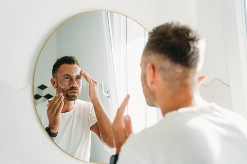 A millennial man is using a face cream in the bathroom. Man's care beauty routine.