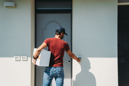 A delivery man is standing in front of the customer's entrance door. He's holding a cardboard box. He's ringing the bell.