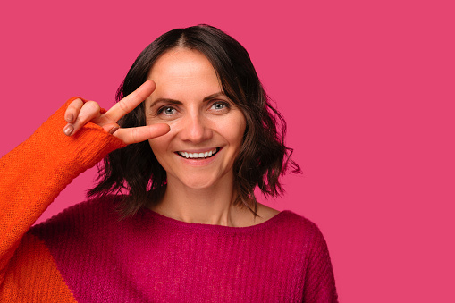 Close up portrait of a smiling mid age woman holding v gesture near her eye over pink background.