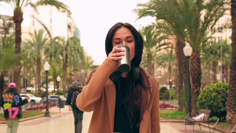 Young woman with headphones walking down the street drinking coffee