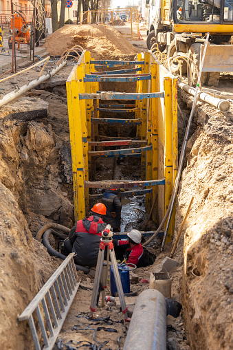 Excavator filling deep excavation supported by trench box with pipe bedding pea gravel during installation of drainage pipe. Reconstruction of technical communications on one of the streets of a European city.