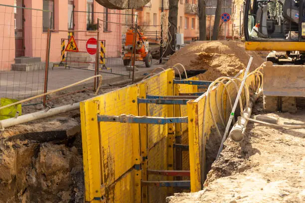Excavator filling deep excavation supported by trench box with pipe bedding pea gravel during installation of drainage pipe. Reconstruction of technical communications on one of the streets of a European city.