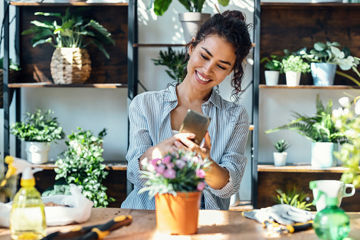 Shot of beautiful smiling woman taking photos with smartphone to plants and flowers in a greenhouse