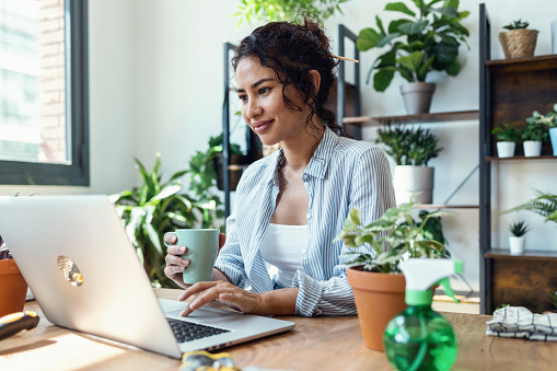 Shot of smiling woman drinking a cup of coffee while working with her laptop in greenhouse.