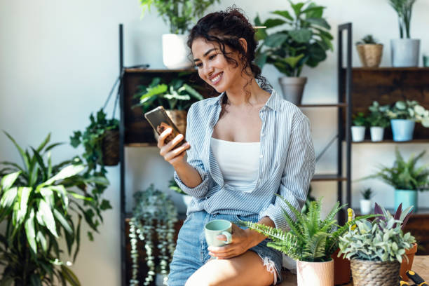 istock Smiling woman drinking a cup of coffee while using her mobile phone in greenhouse. 1471031781