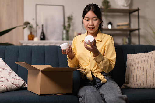 Beauty Shopping. Portrait of happy Asian woman received package, unpacking cardboard beauty box with cosmetics product, holding jars with skincare cream in hands sitting on the sofa couch at home