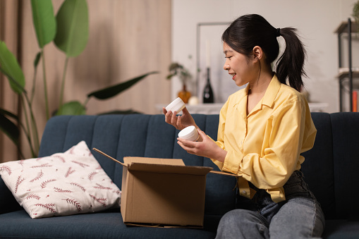 Happy Asian Lady Holding Delivered Moisturizer Jars Unpacking Cardboard Box After Successful Beauty Shopping Sitting On Sofa At Home. Cosmetology Products, Delivery Service Concept