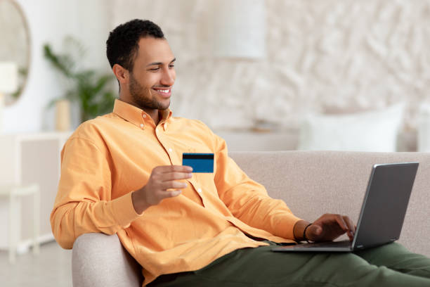 istock Smiling man using pc and credit card sitting on couch 1471032297